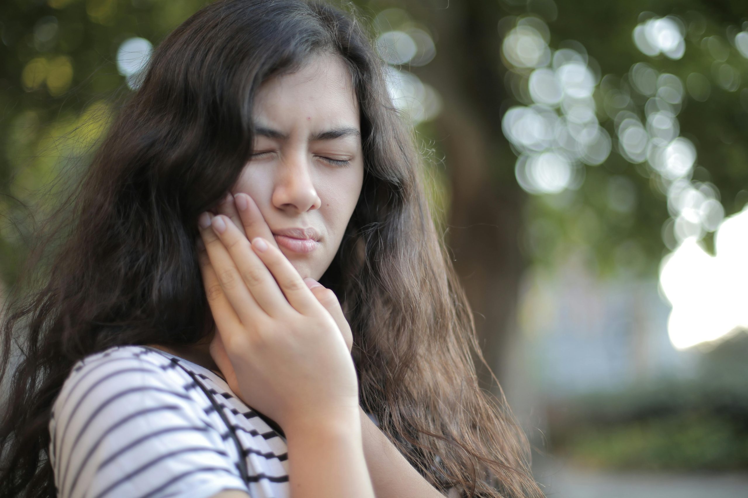 Photo of a young brunette woman with wavy hair holding her hands to her jaw as if in pain.